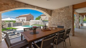 Ein überdachter Essbereich im Freien mit Pool, Steinmauern und Blick auf ein Gebäude.