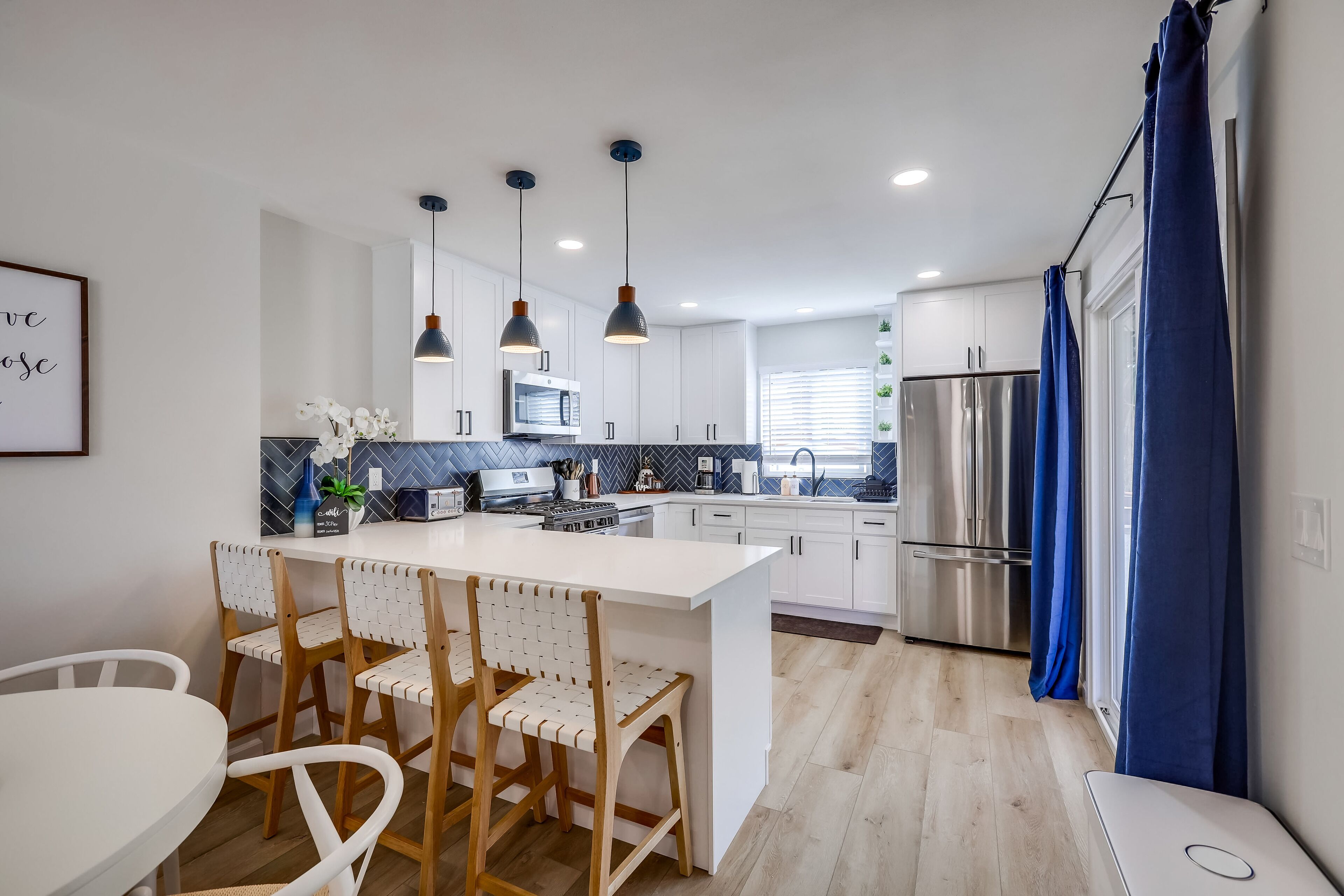 A modern kitchen with white cabinets, a central island, pendant lighting, and a stainless steel refrigerator.