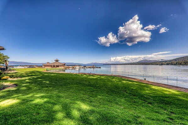 A lakeside view with a grassy area, a wooden dock, and a building with a tower. - Fleur Du Lac 16 By Tahoe Getaways - Lakefront Monthly Rental with Pool, Tennis, Gym, Boat Slip (Homewood)