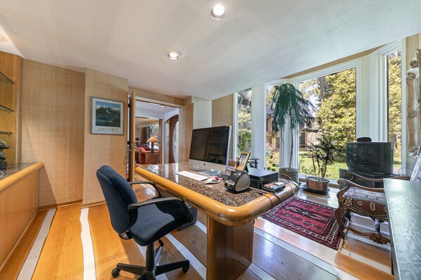Un despacho en casa con escritorio, computadora y silla. Hay un ventanal con vistas a árboles y césped.