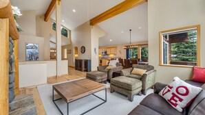 A spacious living area with a wooden coffee table, comfortable seating, a kitchen in the background, and a large window with a view of greenery.