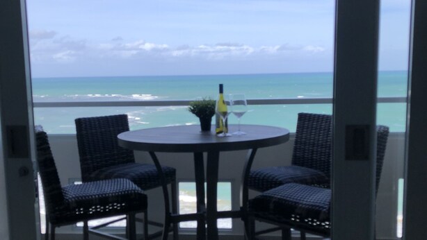 A balcony with wicker chairs and a table, overlooking a beach with the ocean and a cloudy sky. - << NEW >> Completely Renovated, Playa Azul-Tower 1, 17th Floor, 1 Bedroom 1Bath  (Luquillo)