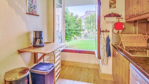 Una cocina pequeña con una mesa de madera, una cafetera, un cubo de basura y vistas a un jardín verde a través de una puerta abierta.