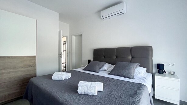 A modern hotel room with a large bed, grey bedding, and a white nightstand with a lamp.