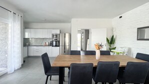 A modern kitchen with white cabinets and stainless steel appliances, a dining area with a wooden table and black chairs, and a white brick wall.