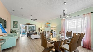 A spacious living area with a glass-top dining table surrounded by brown chairs, a light blue sofa, and a coffee table. There is a ceiling fan, a large window with sheer curtains, and a view of the outdoors.