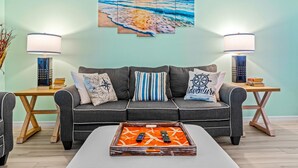A living room with a grey sofa, two side tables with lamps, a tray with remotes, and a wall art of a beach scene.