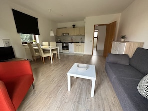 Uma sala de estar moderna com uma poltrona vermelha, um sofá cinza e uma mesa de centro branca. Uma cozinha com armários de madeira clara e uma área de jantar com mesa e cadeiras são visíveis ao fundo.