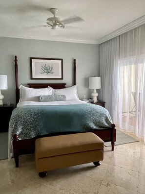 Un dormitorio con una cama grande, un ventilador de techo, un cuadro enmarcado en la pared y una ventana con cortinas translúcidas.