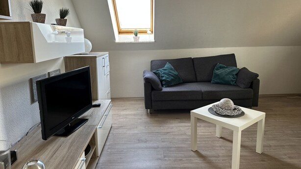Una sala de estar moderna con un televisor de pantalla plana, un sofá gris con cojines turquesa, una mesa de centro blanca y un estante pequeño blanco con plantas en macetas.