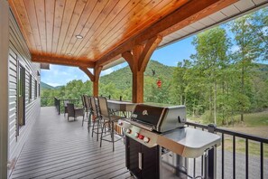 A covered outdoor deck with a grill and seating area, overlooking a forested landscape.