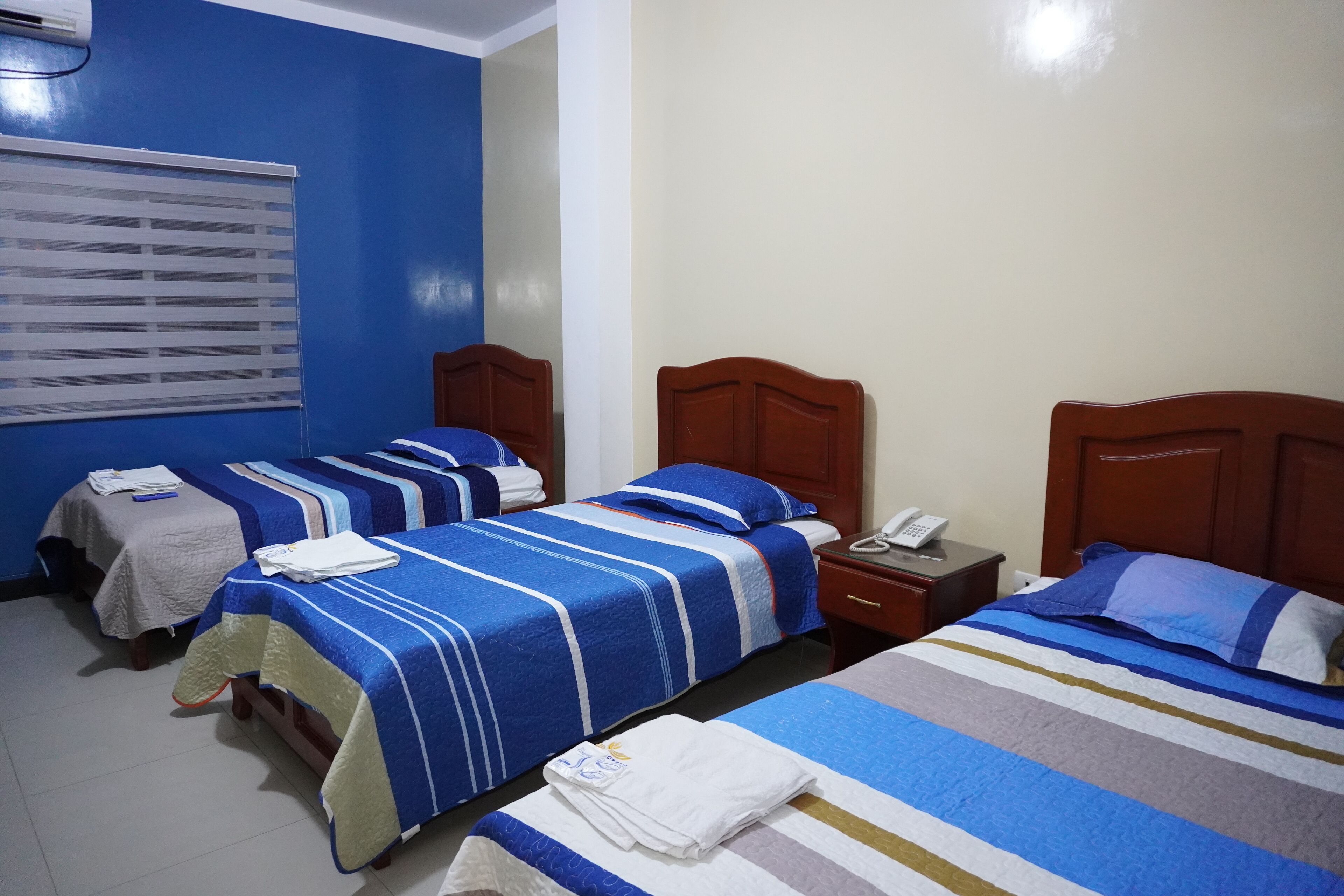Habitación de hotel con tres camas, cada una con ropa de cama azul y blanca a rayas. Se ve un cabecero de madera a la derecha, una mesita de noche con un teléfono, y una ventana con persianas a la izquierda.