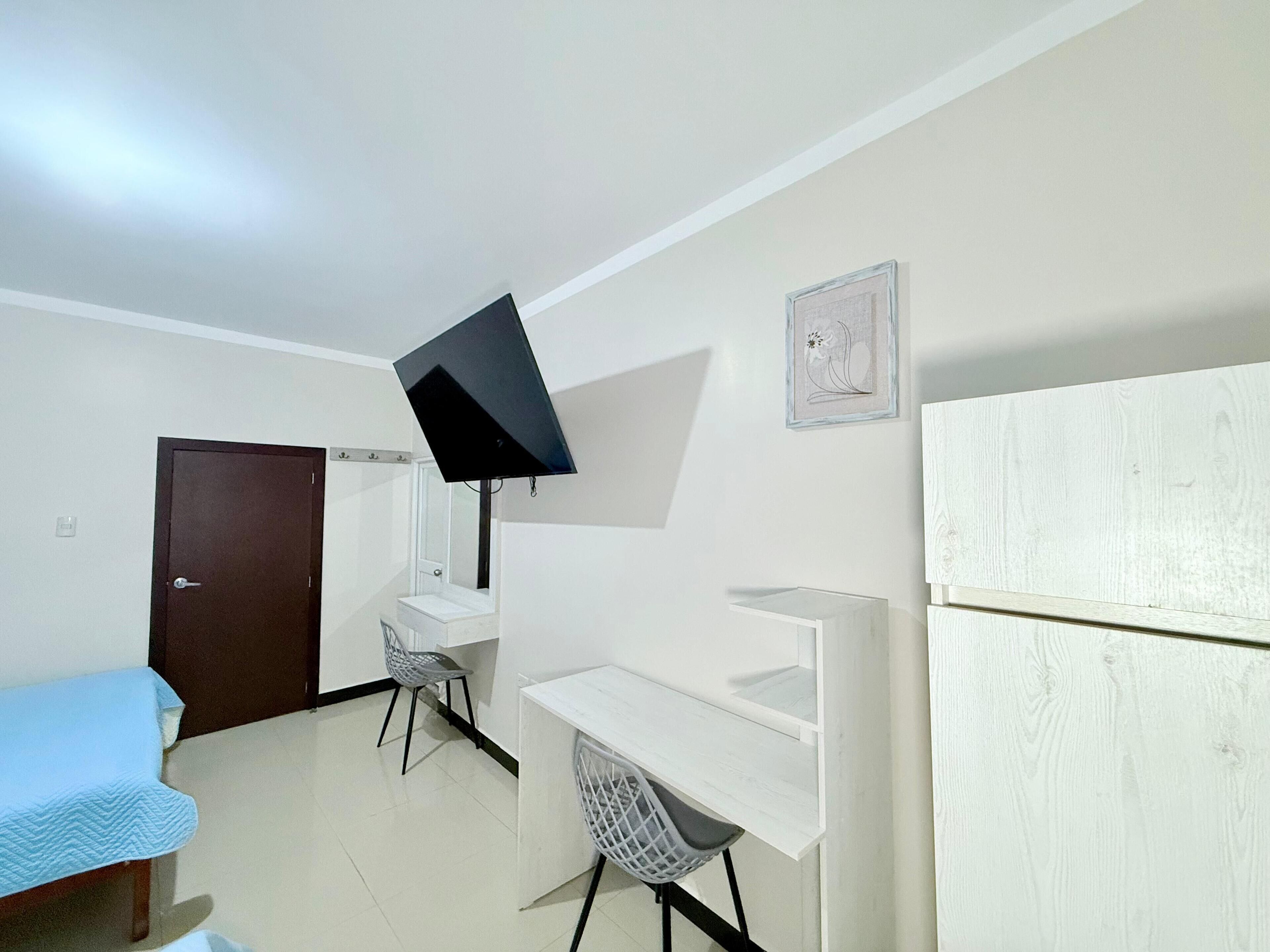 Una habitación pequeña y luminosa con cama, escritorio, silla y televisor montado en la pared.