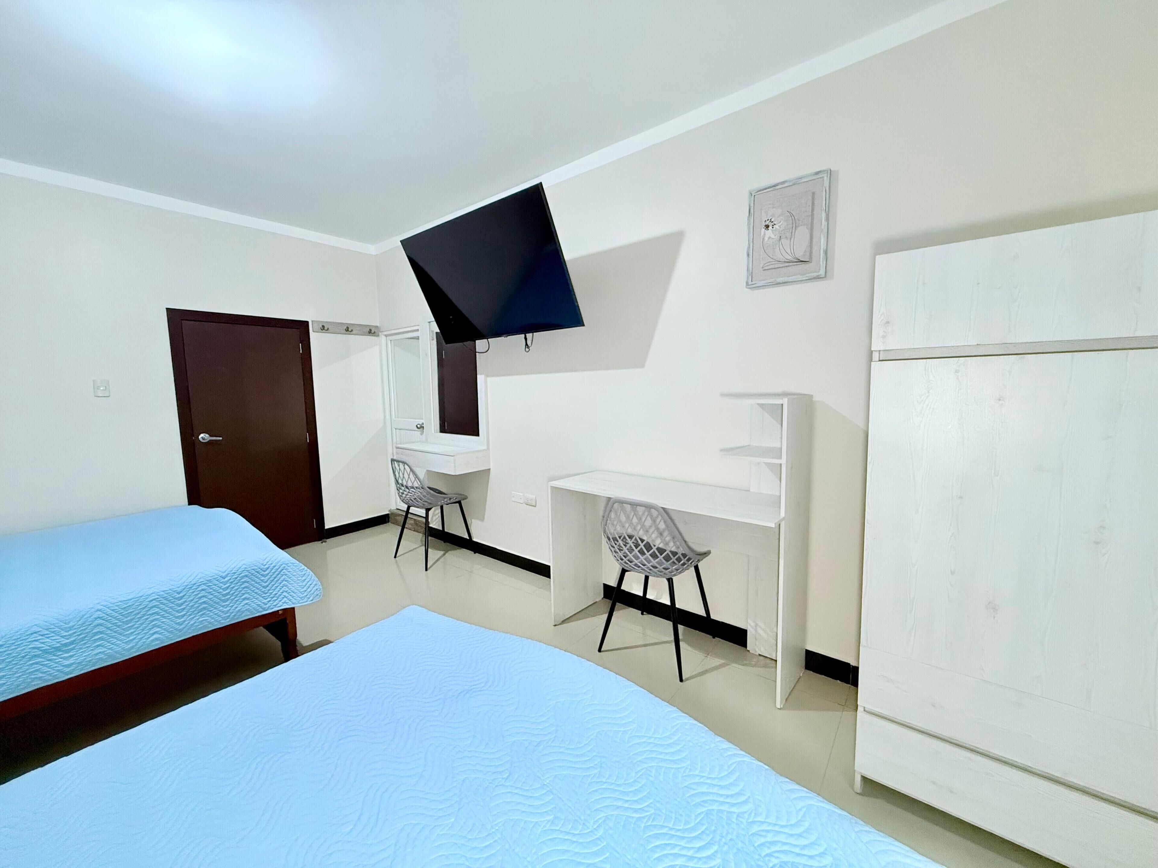 A room with two beds, a desk, a chair, a TV mounted on the wall, and a wardrobe.