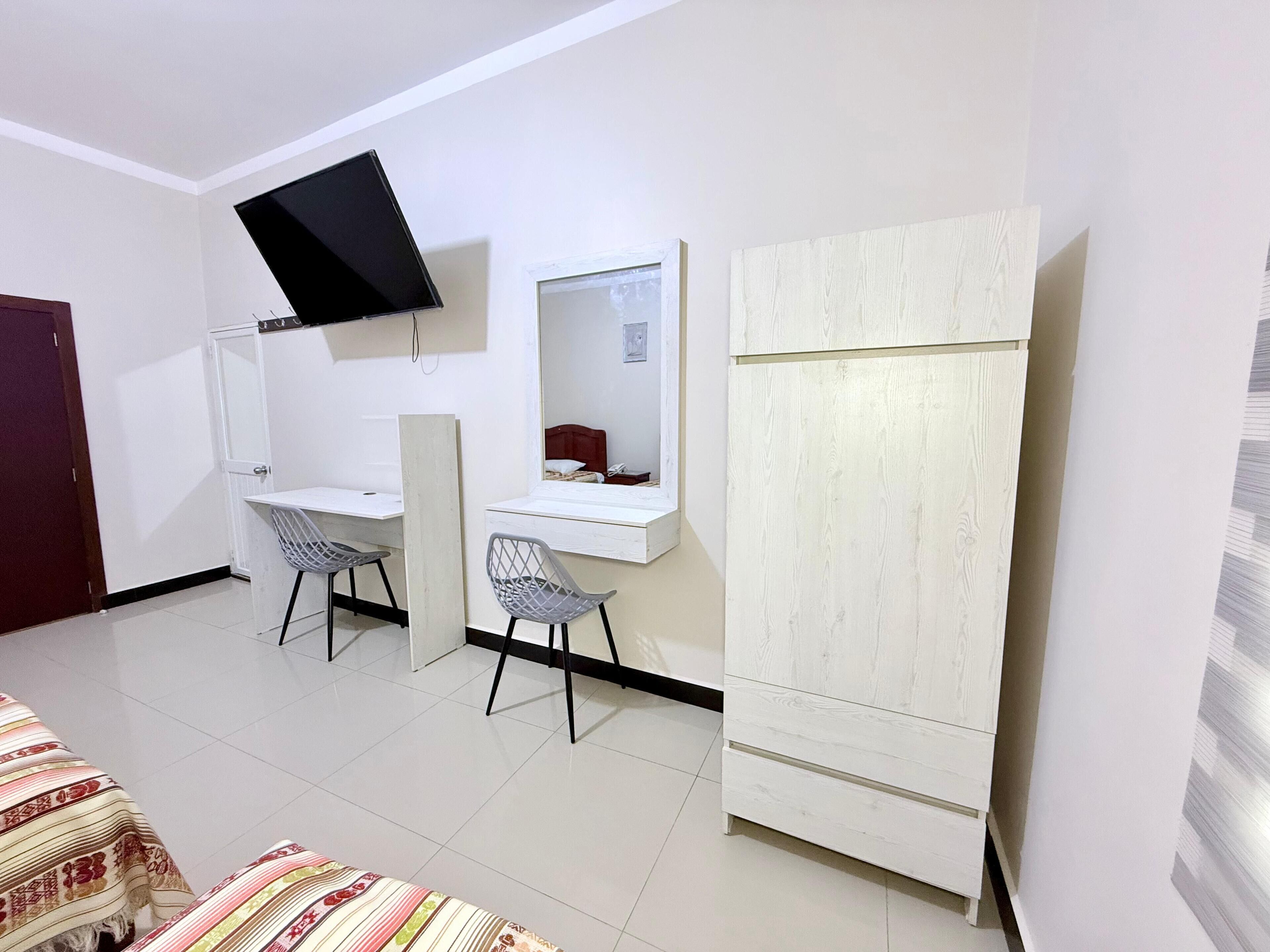 A compact room with a desk, two chairs, a television mounted on the wall, and a wardrobe.
