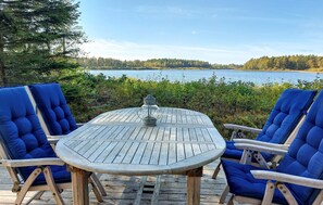 A wooden deck with a round table and four blue cushioned chairs facing a lake.