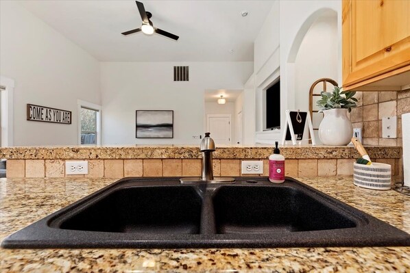 A kitchen with a double sink, granite countertop, a ceiling fan, and a 'COME AS YOU ARE' sign on the wall. - Meridian Patio Home with Games, Seasonal Pool, Close to EVERYTHING! Pet friendly (Meridian)