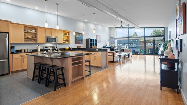 Eine moderne Küche mit Holzschränken, einer zentralen Kochinsel mit Barhocker, Edelstahlgeräten, Pendelleuchten und Blick auf ein Café durch große Fenster.