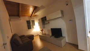 Un soggiorno con un divano, una televisione su un mobile, una finestra e un soffitto in legno.