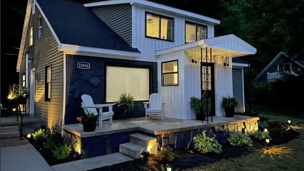 A modern two-story house with a garage, a covered porch, and a well-lit front yard with a small water feature and potted plants.