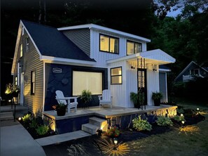 A modern two-story house with a garage, a covered porch, and a well-lit front yard with a small water feature and potted plants. - Remodeled Traverse City Home - Lake View-Golf and Wineries STR #2026-52 (Traverse City)