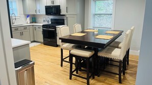 A modern kitchen with white cabinets, a black microwave, and a stove. There is a dining table with chairs and a trash bin.