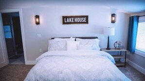 A bedroom with a bed, a wall-mounted sign reading "LAKE HOUSE", two wall sconces, a bedside table with a lamp, and a window with curtains.
