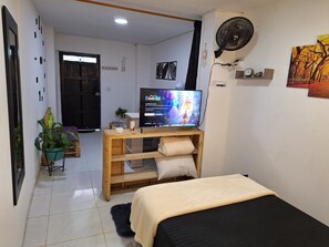 A bedroom with a bed, a TV on a wooden stand, a fan, and a small shelf with decorative items.