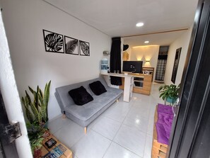A modern living room with a grey sofa, black cushions, and a coffee table. There is a TV on a wooden stand, a small kitchen area with a fridge and a sink, and a potted plant. - OSIMiRI apartamentos (San Rafael)