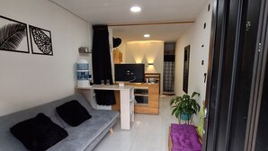 Un ambiente compatto con un divano grigio, una piccola panca in legno, un distributore d'acqua e un televisore.