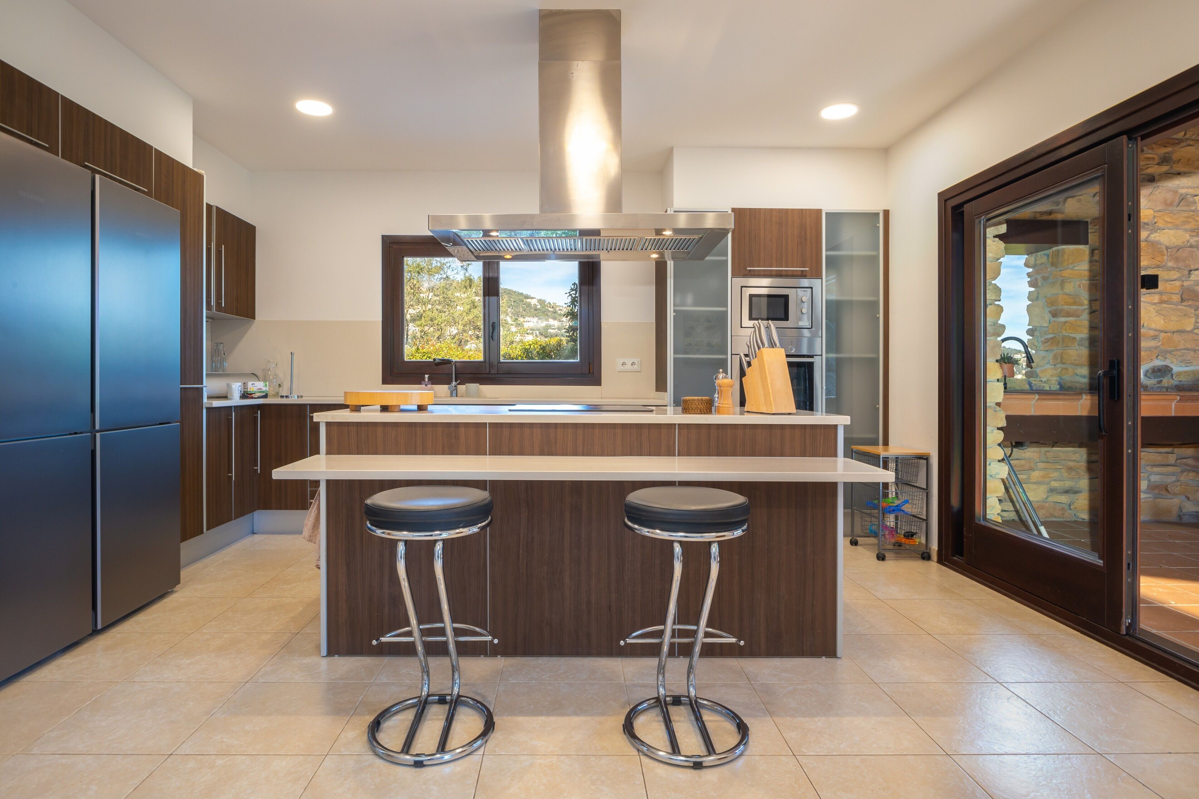 A modern kitchen with a central island, two bar stools, stainless steel appliances, and a view of the outdoors.