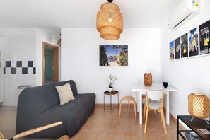 A modern living room with a sofa, a dining table with chairs, a small side table with a plant, and a wall-mounted air conditioner.