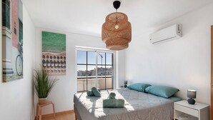 Un dormitorio con una cama, un ventanal, una planta en maceta, aire acondicionado instalado en la pared y una lámpara colgante.