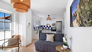 Una cocina moderna con armarios blancos, una zona de comedor con sofá y sillón, y un ventanal grande.