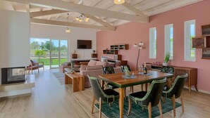 A spacious living area with a high ceiling, wooden beams, a fireplace, a dining table with chairs, a sofa set, and a television mounted on the wall.