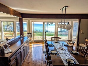 A spacious dining area with a long table set for six, large windows with a view of the outdoors, and a modern kitchen with dark wood cabinetry.