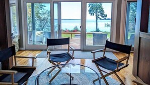 A room with a glass door leading to a deck with a view of the water. There are two foldable chairs and a glass table.