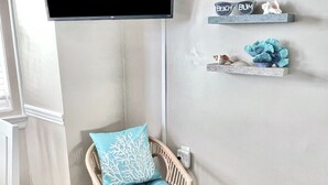A room with a television mounted on the wall displaying 'NETFLIX', a wicker chair with a blue cushion, and decorative shelves with various items. - Ocean City, NJ - Feet to the Boardwalk, New Listing, Turnkey & Clean! (Ocean City)