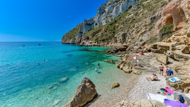 A rocky beach with clear turquoise water, people swimming and sunbathing, and a cliff with vegetation. - Casa Peponi - By Almarina Villas (Javea)