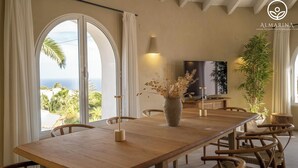 A dining area with a wooden table, chairs, a vase with dried flowers, a TV on a wooden cabinet, and a large window with a view of greenery and the sea. - Casa Peponi - By Almarina Villas (Javea)
