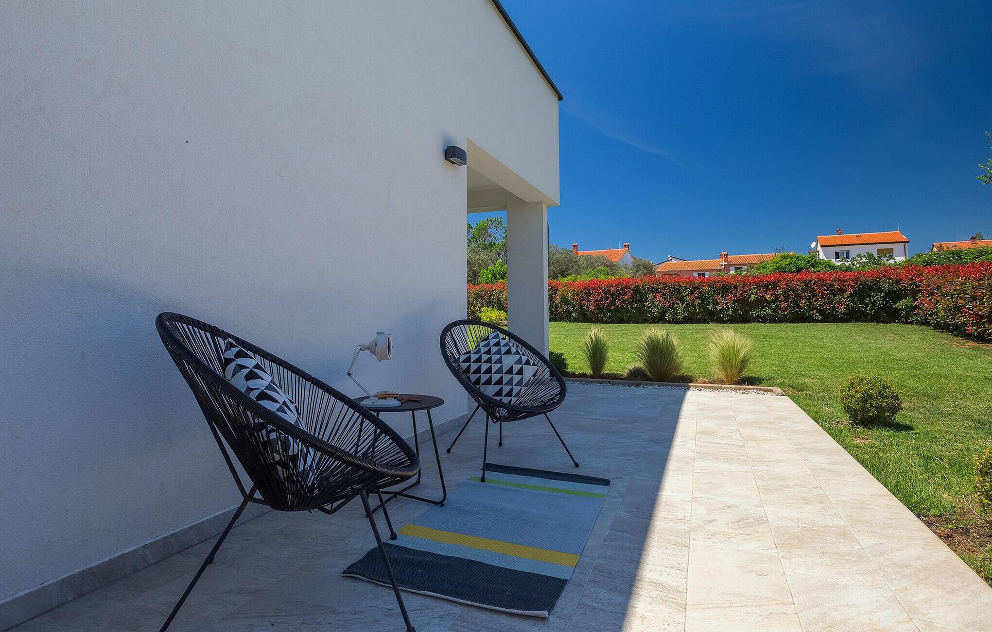 A modern outdoor seating area with two black chairs and a small table, a white wall, and a well-maintained garden with a hedge and red flowers.