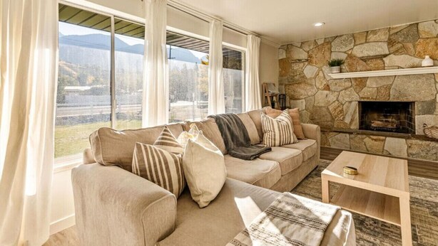 A cozy living room with a stone fireplace, a large window with curtains, and a sofa with multiple cushions. - Canyon View Cottage | Backyard | Free Massage* (Provo)