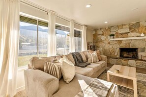 A cozy living room with a stone fireplace, a large window with curtains, and a sofa with multiple cushions. - Canyon View Cottage | Backyard | Free Massage* (Provo)