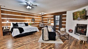 A modern bedroom with a wooden floor, a bed with white bedding, a black sofa, a fireplace, and a TV mounted on the wall. - Honeymoon Getaway•Hot Tub•Fireplace•VIEWS•Seclusion (Sevierville)