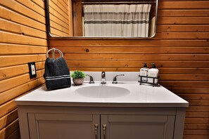 Ein Badezimmer mit Holzvertäfelung, einem weißen Waschbecken, einem Spiegel, einem Handtuch, einer Pflanze und Seifenspendern.