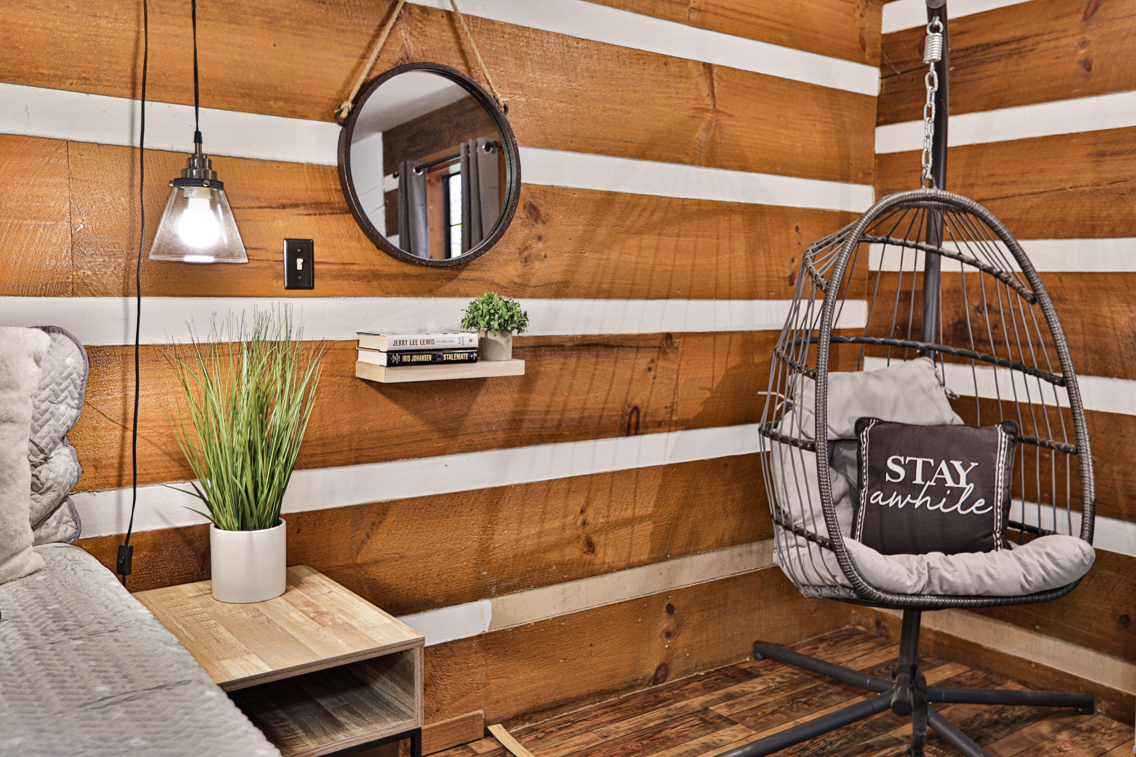 Una habitación acogedora con una silla colgante, un estante pequeño con libros y una planta, una mesita de noche y un muro de madera con tablones horizontales.