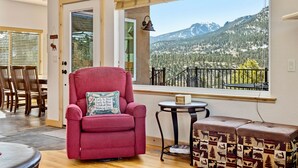 A room with a large window offering a mountain view, a red armchair, a small round table, and a bench with a patterned cushion.