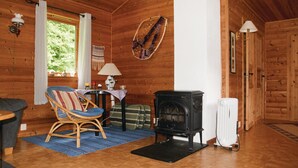 A cozy cabin interior with a wooden floor, a black wood-burning stove, a wicker chair, a table with a lamp, and a window with curtains. - Amazing home in Sandeid with lake view (Sandeid)