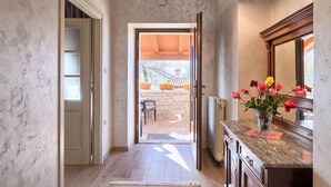 Un couloir doté d’un sol en pierre, d’une porte en bois, donnant sur un patio extérieur agrémenté de plantes en pot, et d’un comptoir en marbre sur lequel est posé un vase de fleurs.