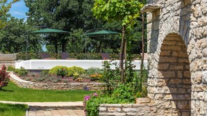 Un arc en pierre mène à un espace extérieur abrité, aménagé avec des tables et recouvert d’une nappe blanche, et est entouré d’un jardin où poussent différentes plantes et fleurs.